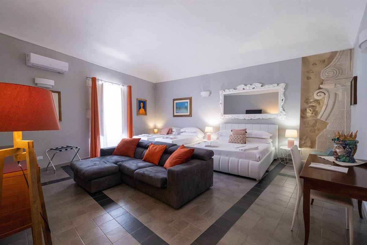 A modern hotel room with a grey sofa, a bed with white bedding, a large mirror, and a small table with a vase.
