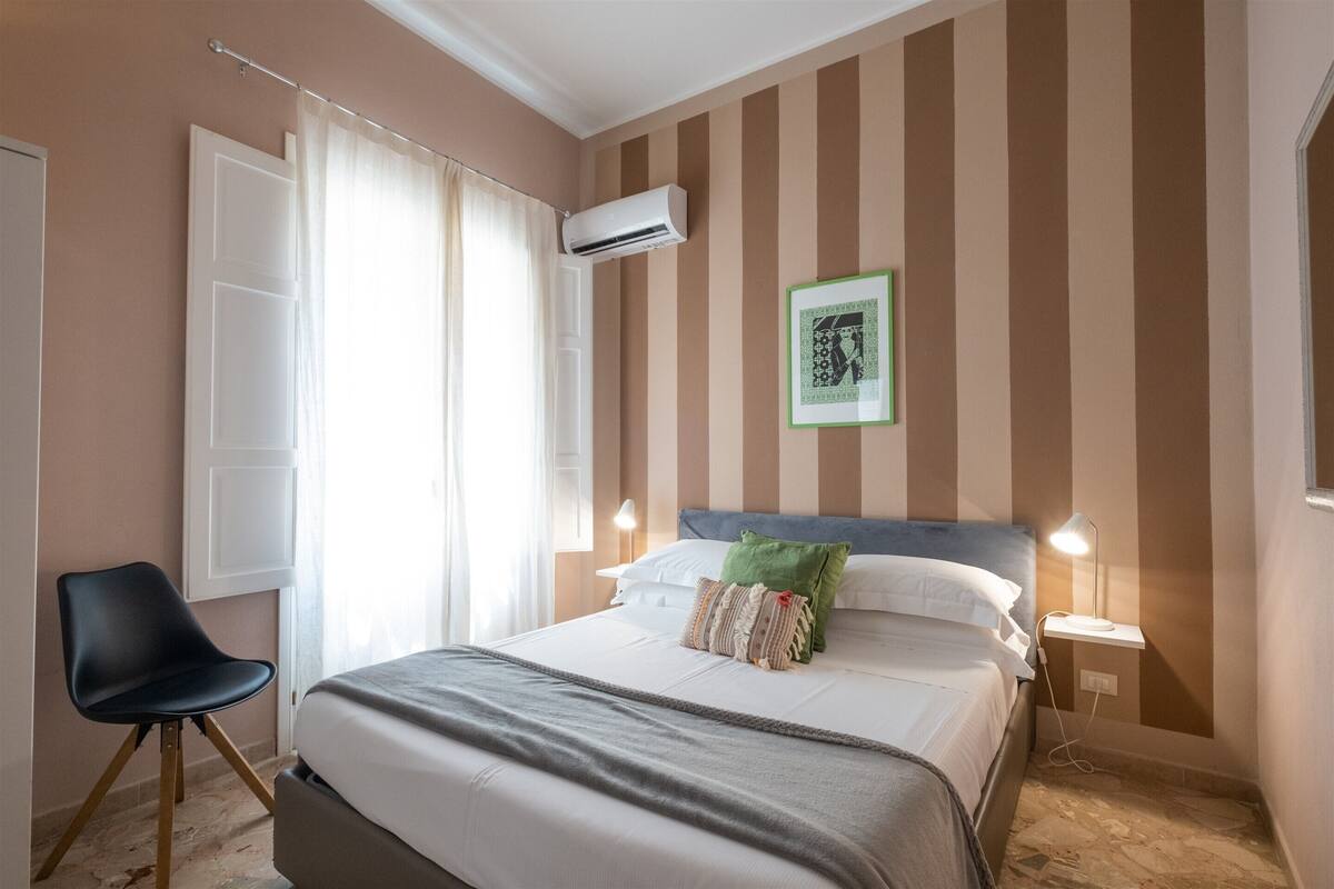 A hotel room with a bed, a chair, an air conditioning unit, a bedside table with a lamp, and a framed picture on the wall.