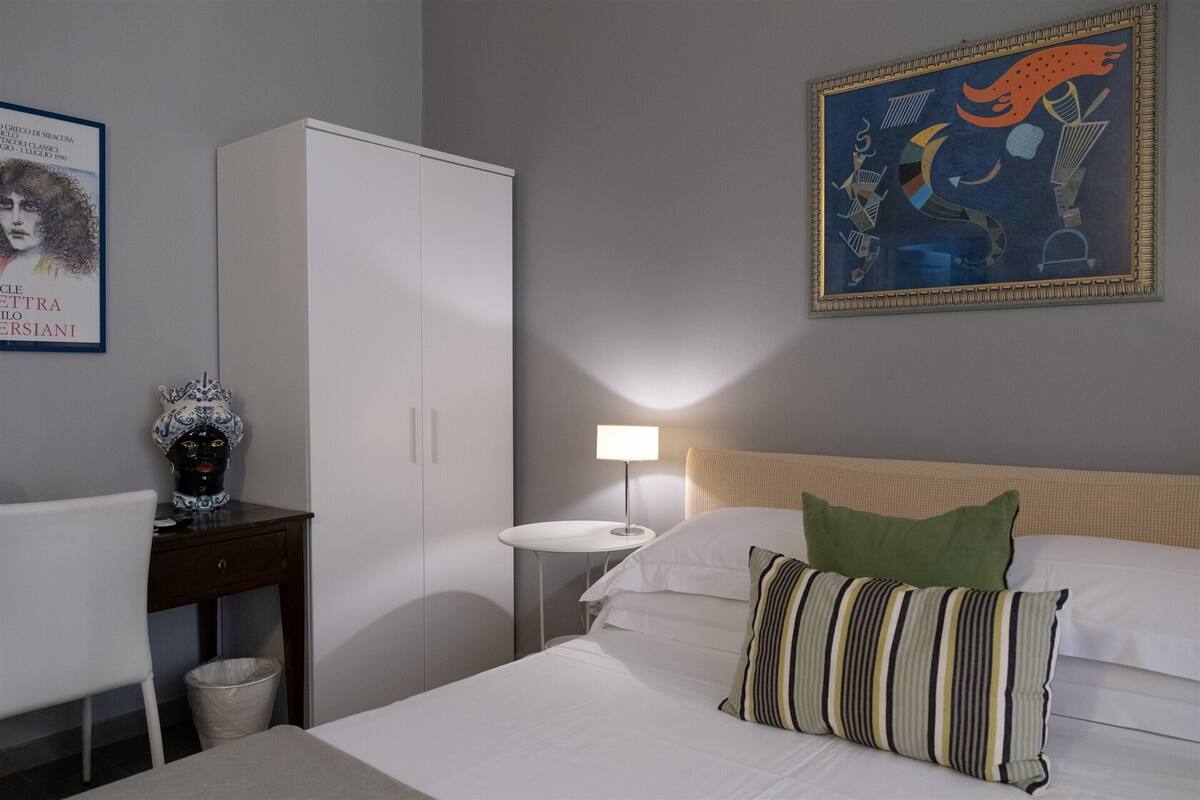 A hotel room with a bed, a desk, a wardrobe, a lamp, and a painting on the wall.