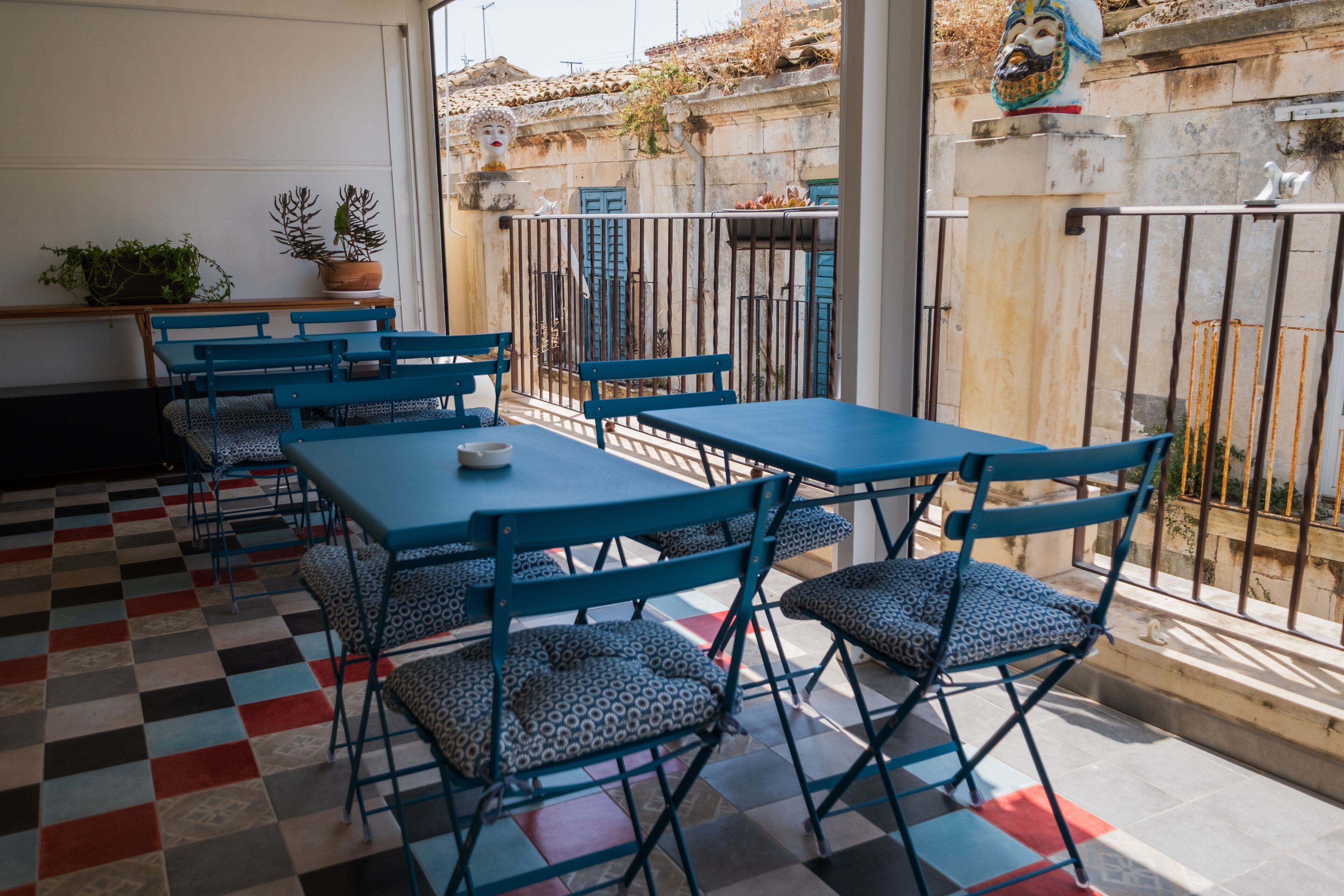 Een terras met blauwe tafels en stoelen, een geruit vloertje en plantenbakken.