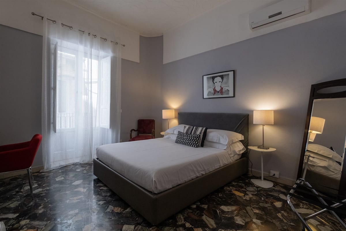 A hotel room with a large bed, two bedside lamps, a red chair, a large mirror, and a window with sheer curtains.