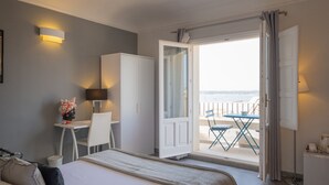 A hotel room with a bed, a desk, a chair, a small table, and a balcony with a view of the sea. - Palazzo Gilistro (Syracuse)