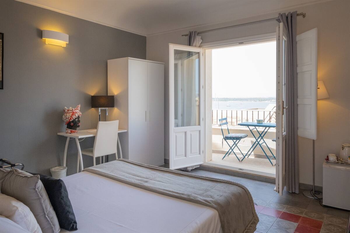 A hotel room with a bed, a desk, a chair, a small table, and a balcony with a view of the sea.