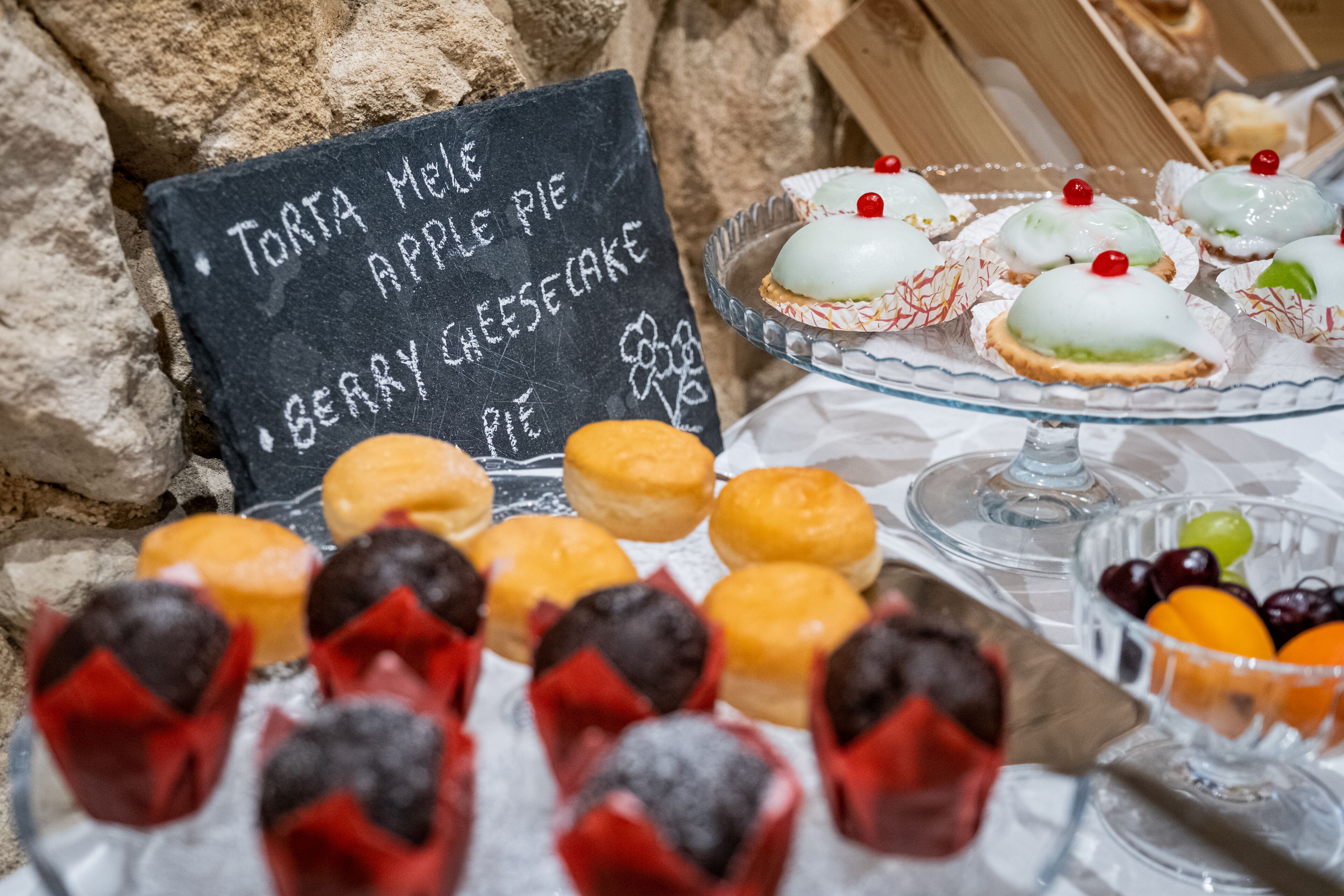 A dessert display with various pastries and a chalkboard sign listing items like Torta Mel, Apple Pie, and Berry Cheesecake.