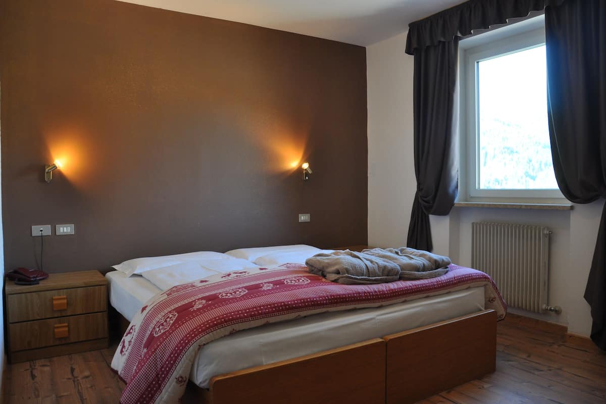 Een slaapkamer met een bed, twee wandlampen, een raam met gordijnen, een radiator en een nachtkastje van hout met lades.