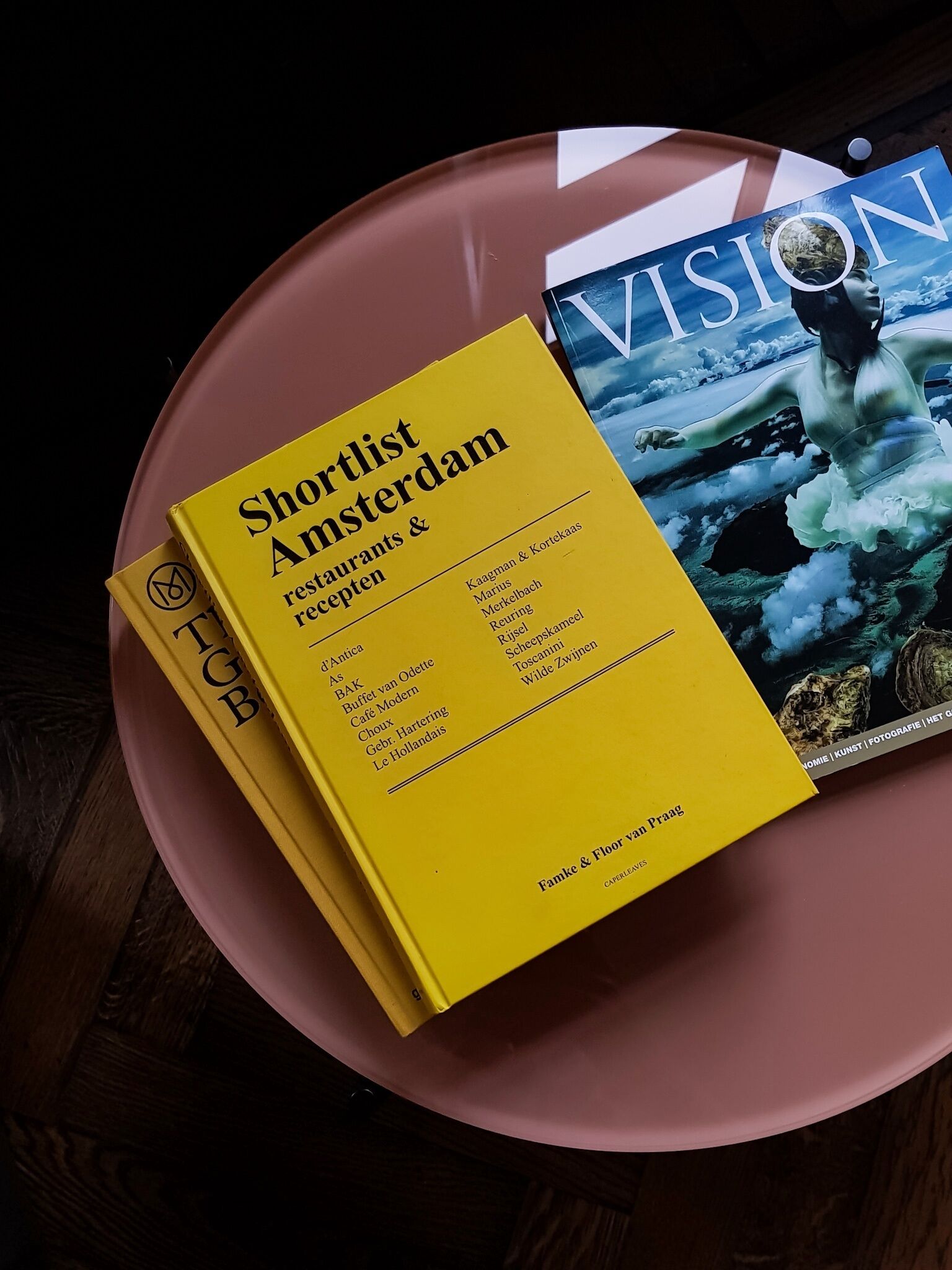Un folleto amarillo con el título 'Shortlist Amsterdam Restaurants & Recepten' sobre una superficie rosa.