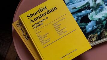 A yellow booklet titled 'Shortlist Amsterdam Restaurants & Recepten' on a pink surface.