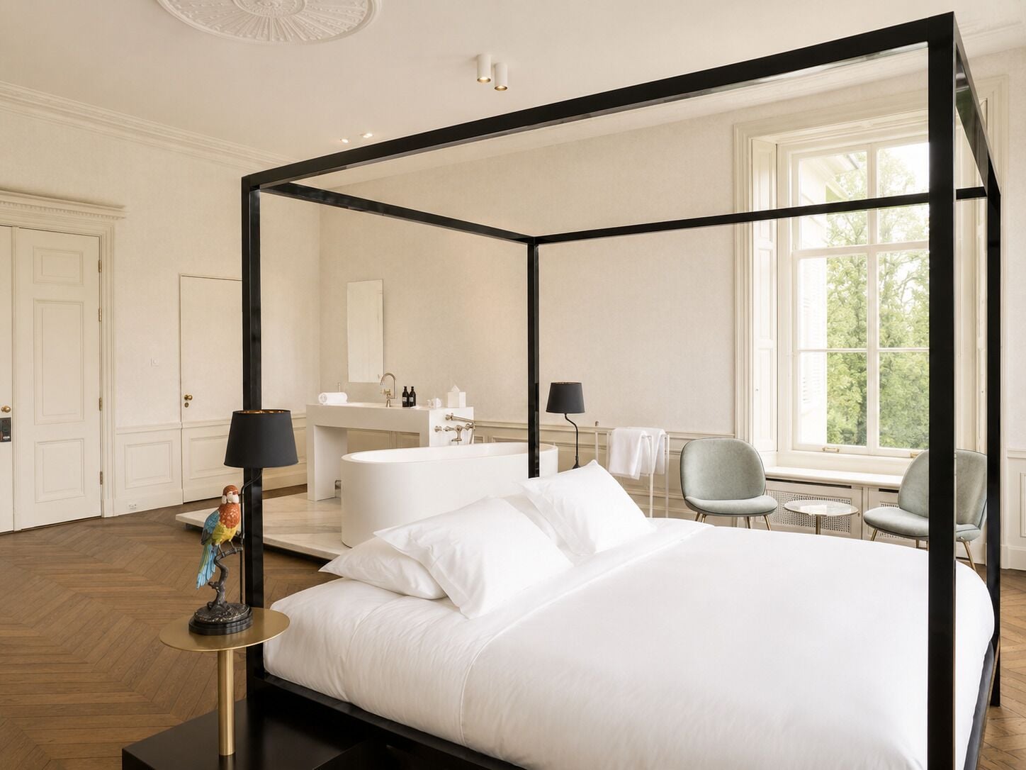 Un lit à baldaquin blanc, une table de chevet ronde avec une lampe, une salle de bain blanche avec un lavabo et un miroir, et une vue sur les arbres à travers une grande fenêtre.