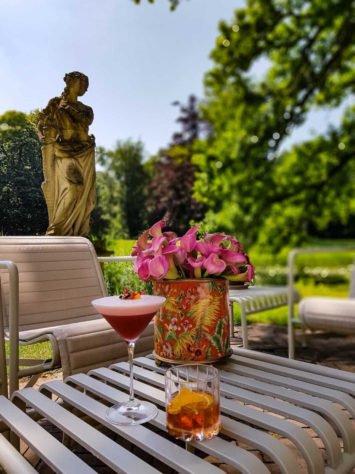 A garden setting with a statue, outdoor seating, and a table with drinks and flowers.