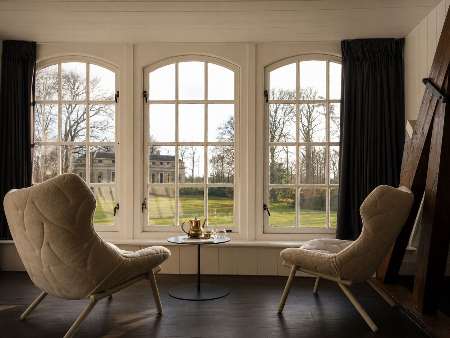 A room with three large arched windows, a small round table with a decorative item, and two upholstered chairs.