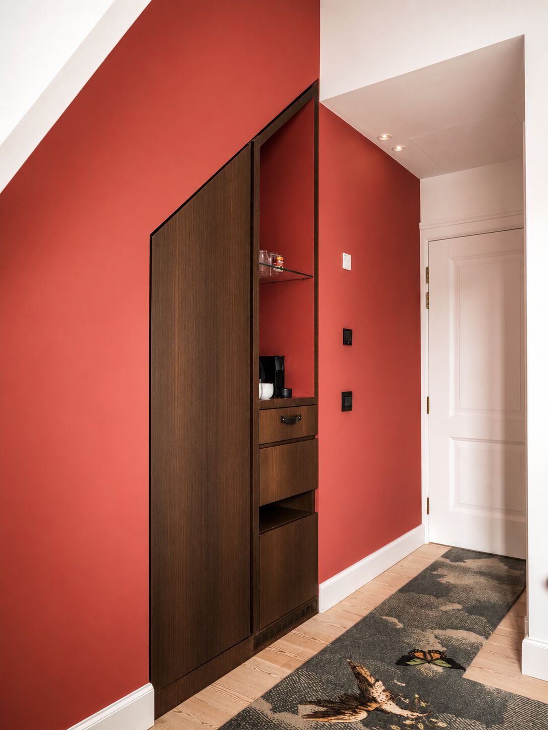 A hallway with a red wall, a wooden wardrobe, a white door, and a patterned carpet.