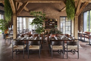 Restaurante com vigas de madeira, janelas amplas e plantas penduradas. Mesas montadas com copos de vinho e pratos.