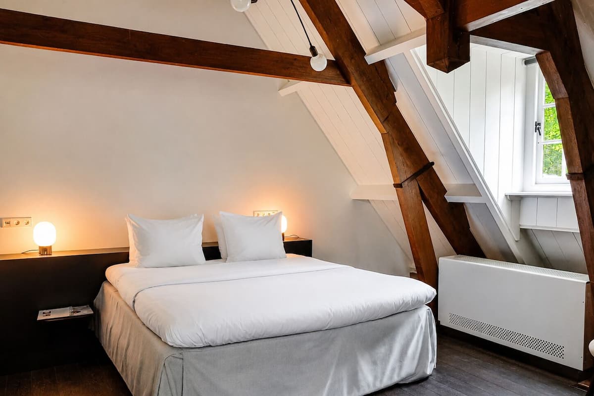 Ein Schlafzimmer mit schrägverdeckter Decke, einem Bett mit weißer Bettwäsche, zwei Nachttischlampen und einem Fenster mit Blick ins Grüne.