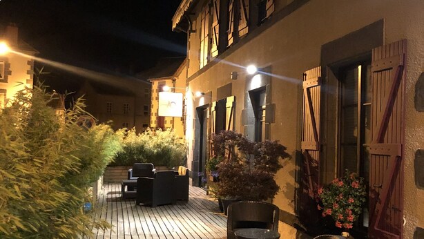 A wooden deck with outdoor seating, surrounded by buildings with traditional architecture and illuminated by ambient lighting. - A l'Aube des Volcans (Charbonnières-les-Vieilles)