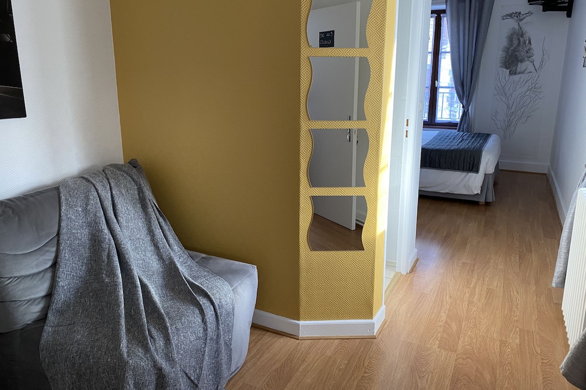 A modern living room with a sofa, a small table, and a bed in the background.