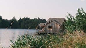 En træhytte med terrasse, beliggende ved en sø med træer i baggrunden.