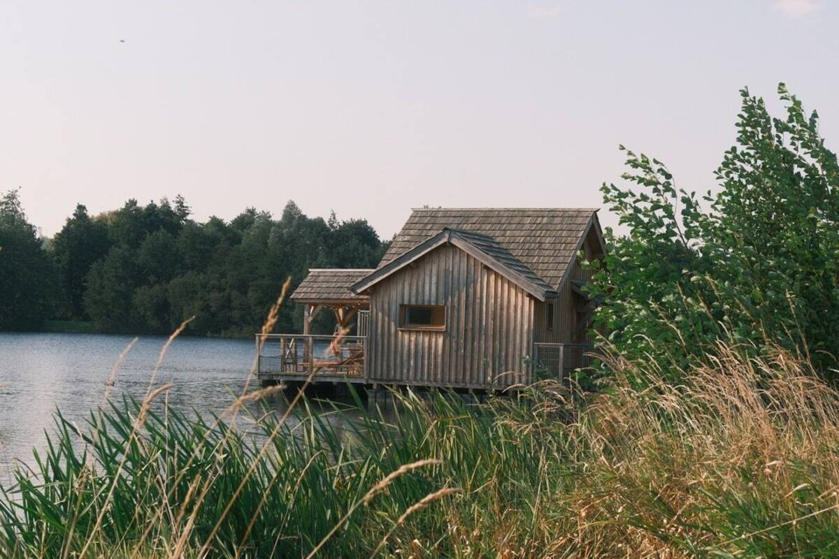 A wooden cabin with a deck, situated by a lake with trees in the background.