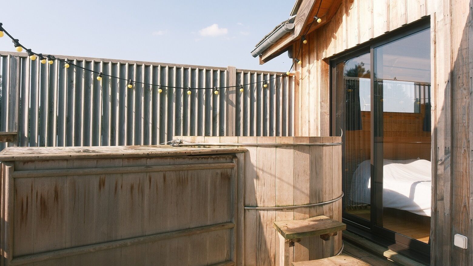 Ein Holzgebäude mit einer großen Schiebeglastür, die zu einem Bett, einem Holzfass und einem Wellblechzaun führt.
