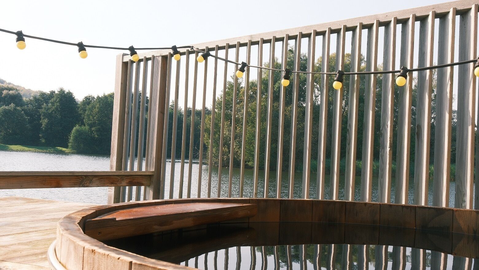 Een houten terras met een hottub, een lichtslingering en uitzicht op een meer en bos.