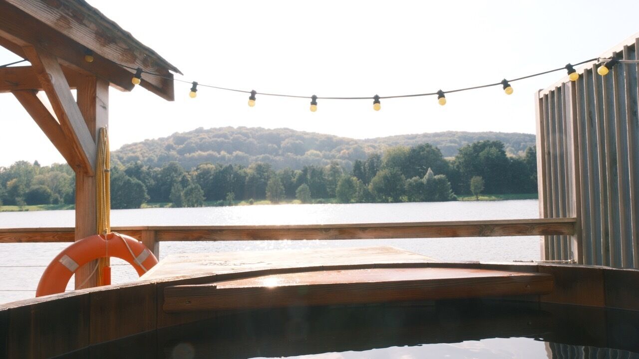 Une terrasse en bois avec un jacuzzi, des guirlandes lumineuses et une vue sur un lac et une forêt.