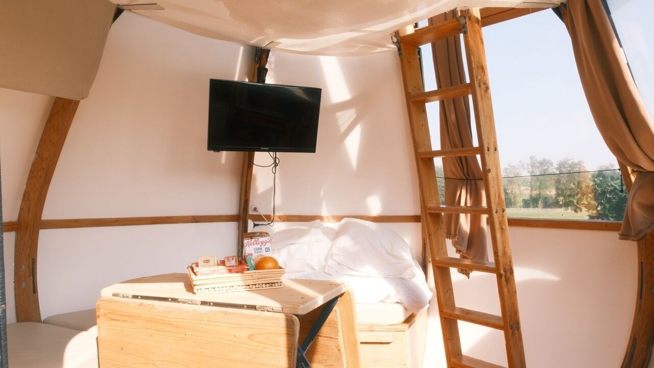 Una habitación acogedora con cama, una mesita con objetos, televisor montado en la pared y ventana con cortinas.