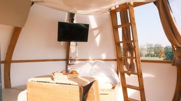 Una habitación acogedora con cama, una mesita con objetos, televisor montado en la pared y ventana con cortinas.