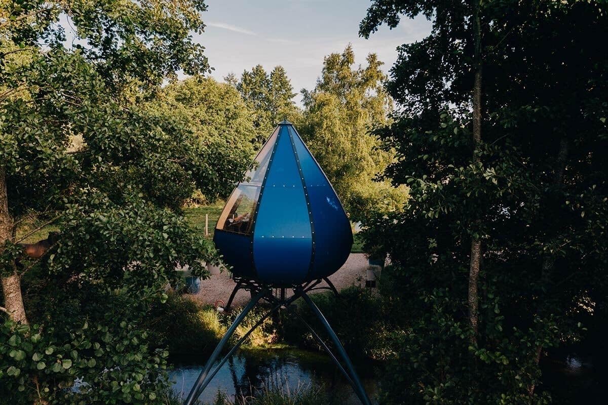 A blue, pyramid-shaped structure in a park with trees and a body of water.