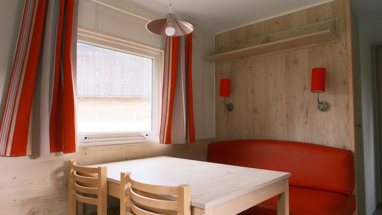 Una habitación con una mesa de madera, dos sillas, un sofá rojo, una ventana con cortinas y dos lámparas fijadas a la pared.
