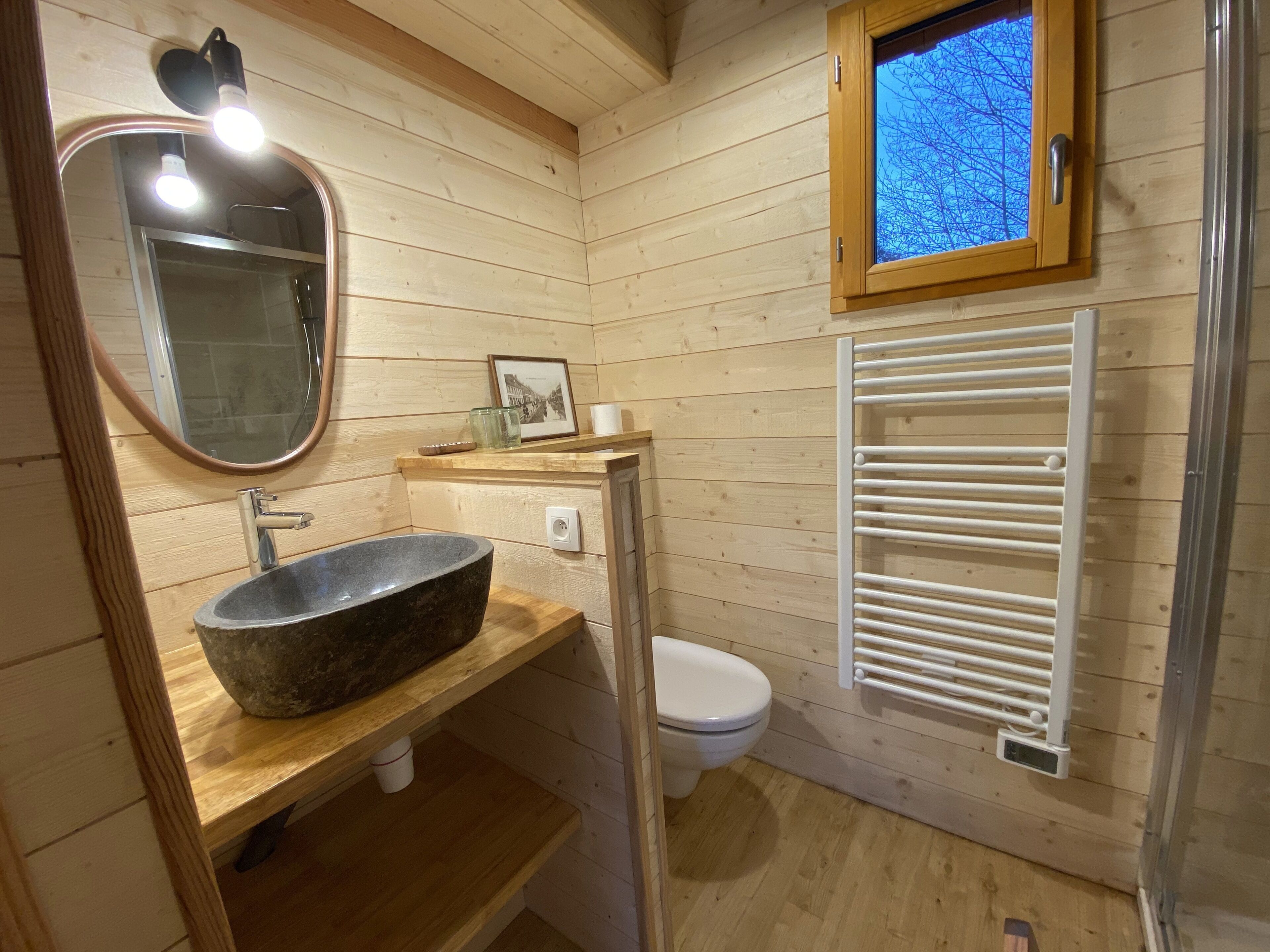 A wooden-paneled bathroom with a stone sink, a white toilet, a mirror, and a window with a view of trees.