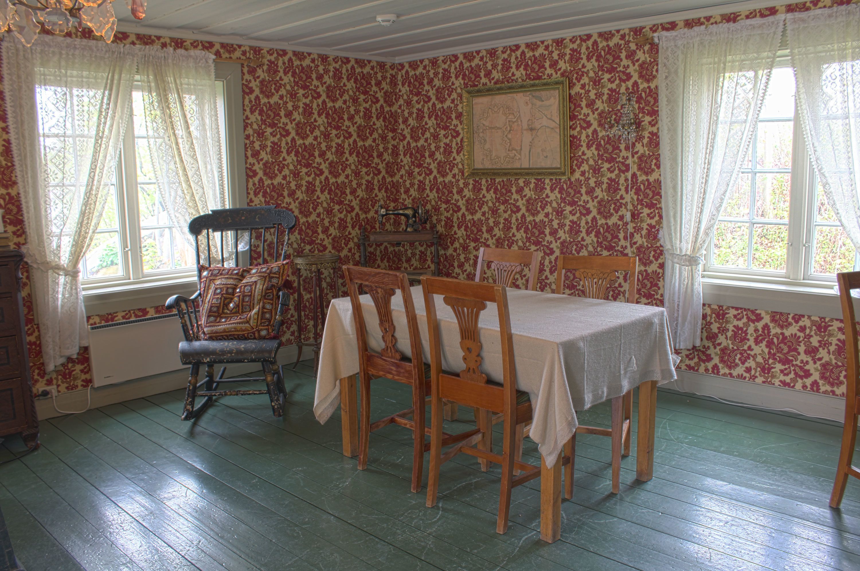 Une salle à manger avec une table dressée pour quatre personnes, entourée de chaises en bois, d’un fauteuil à bascule et d’une machine à coudre ancienne.