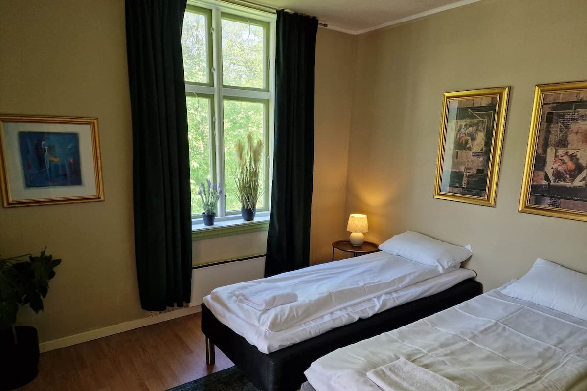 Habitación de hotel con dos camas, una ventana con cortinas, una lámpara en una mesita de noche y cuadros enmarcados en la pared.