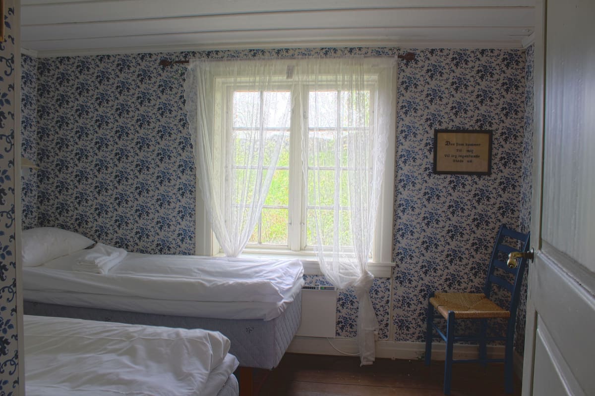 Una habitación con dos camas, una ventana con cortinas translúcidas, una silla y una pared con estampado floral.