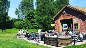An outdoor seating area with tables and chairs arranged in rows, a brick building with a sloped roof, and a grassy area with trees in the background. - Magasinet (Fredrikstad)