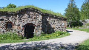 A stone building with a black door and arched windows, surrounded by greenery and a dirt path. - Magasinet (Fredrikstad)