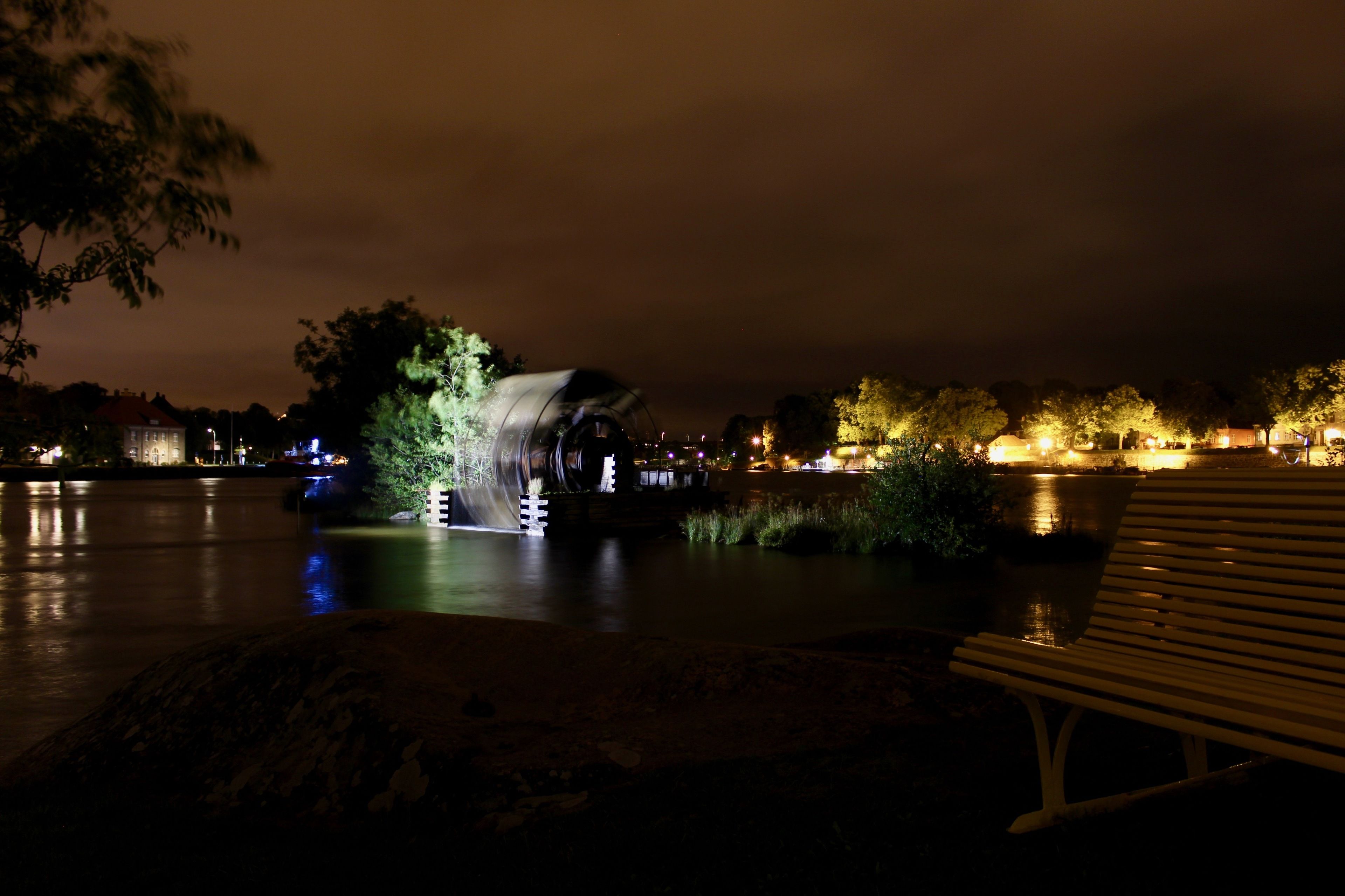A nighttime view of a park with a water feature, a bench, and illuminated trees.