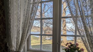 A window with a view of a park and a body of water, a potted plant with flowers, and a lace curtain. - Magasinet (Fredrikstad)