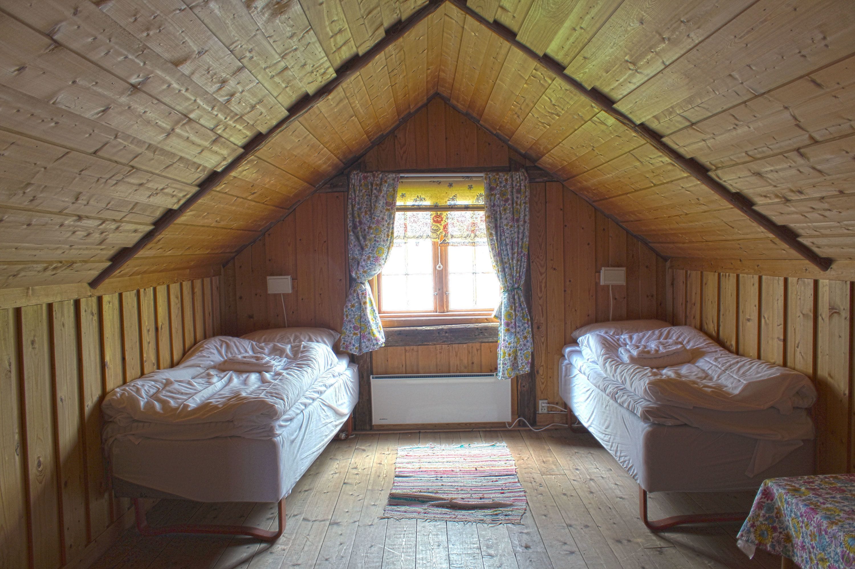 Un pequeño dormitorio en el ático con dos camas individuales, una ventana con cortinas florales y un techo inclinado de madera.