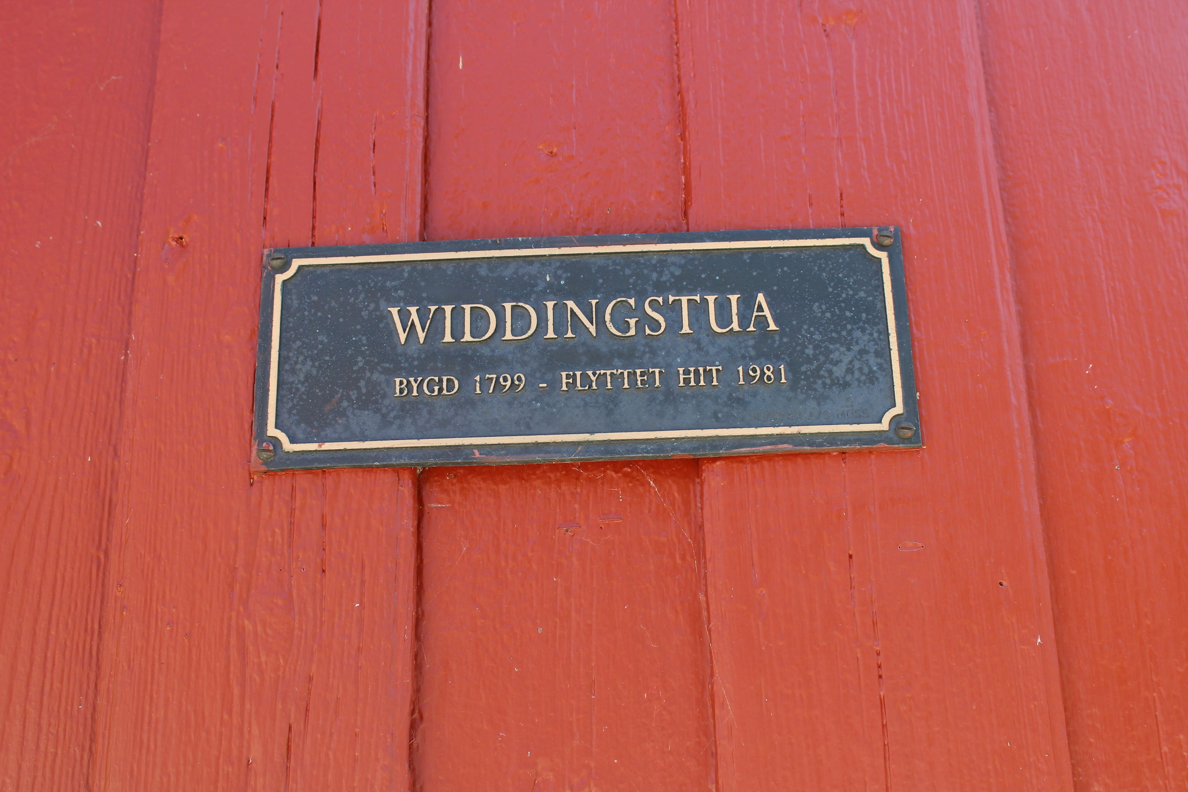 A plaque on a red wooden surface with the text "WIDDINGSTUA BYGD 1799 - FLYTTET HIT 1981".