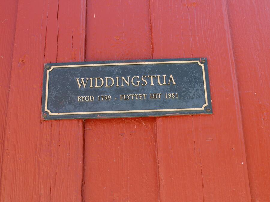 A plaque on a red wooden surface with the text "WIDDINGSTUA BYGD 1799 - FLYTTET HIT 1981".