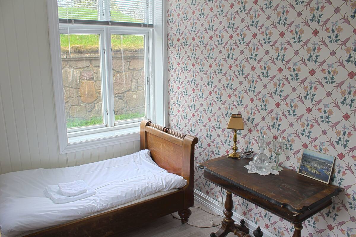 Una habitación con una cama individual, una mesita de noche de madera, un ventanal con vistas a la vegetación y papel tapiz floral.