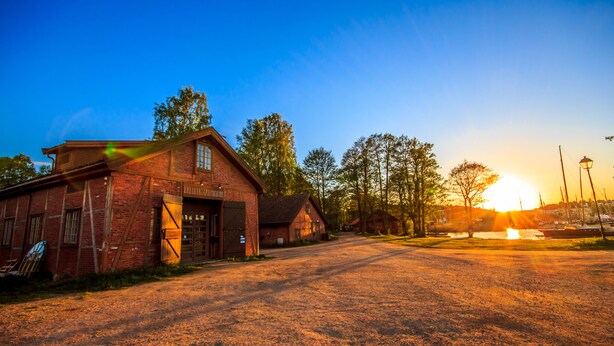A brick building with a garage door, a dirt road, and trees in the background. - Magasinet (Fredrikstad)