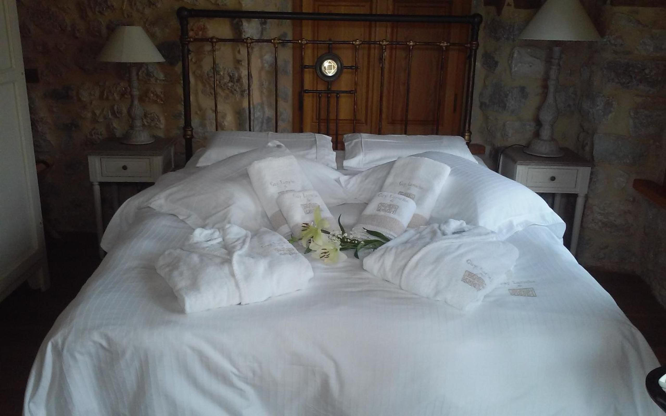 Una cama bien hecha con ropa de cama y toallas blancas, un arreglo floral decorativo y un cabecero de madera.