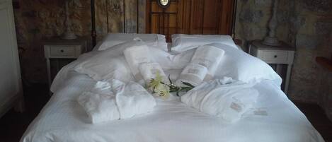 Una cama bien hecha con ropa de cama y toallas blancas, un arreglo floral decorativo y un cabecero de madera.
