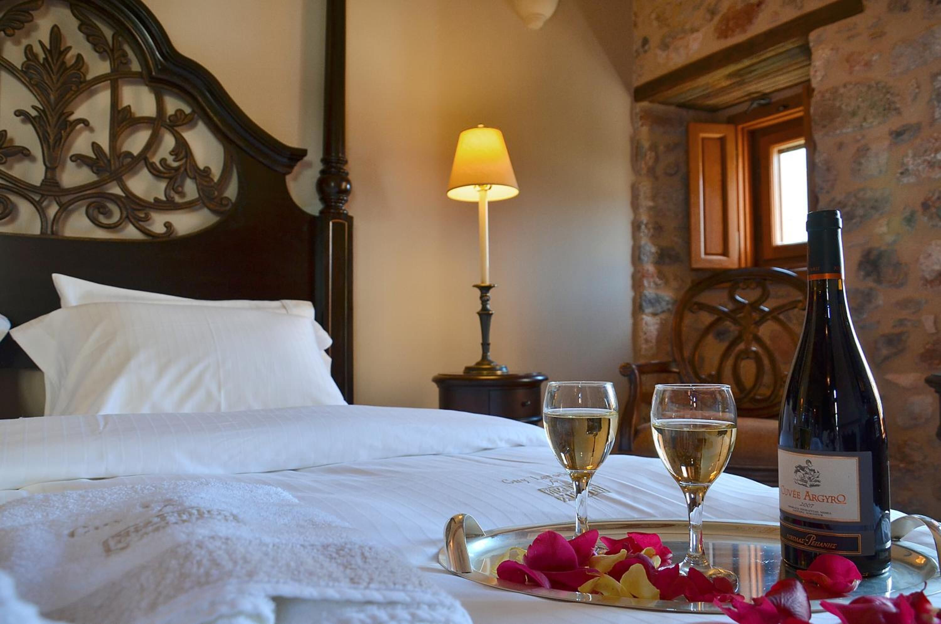 A neatly made bed with white linens, a tray with rose petals and two glasses of white wine, and a bottle of wine on a table.