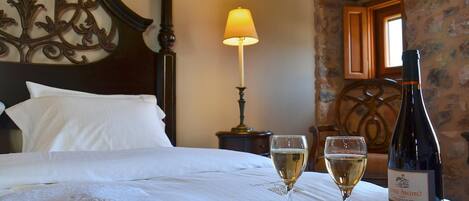 A neatly made bed with white linens, a tray with rose petals and two glasses of white wine, and a bottle of wine on a table.