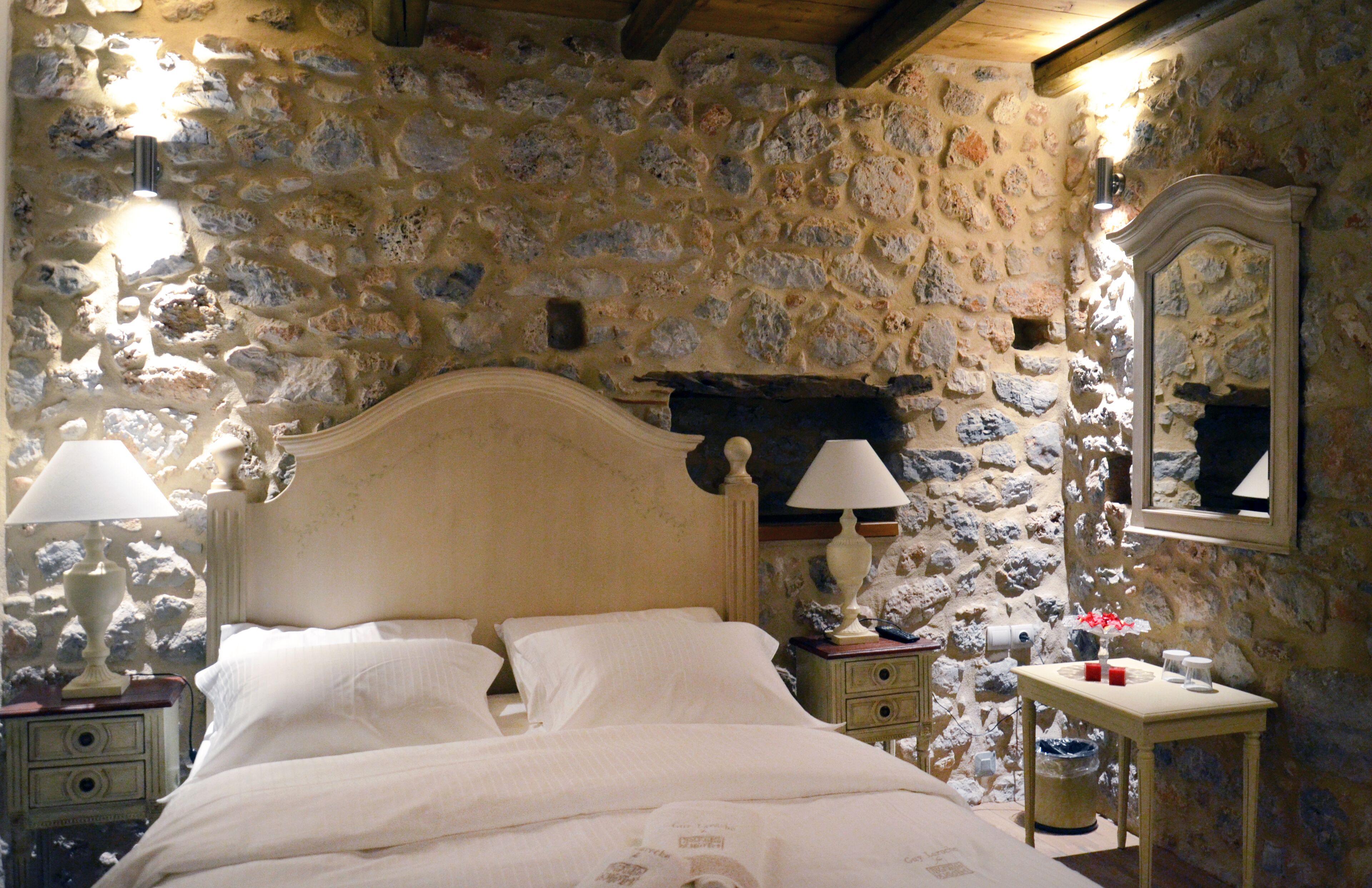 A bedroom with a stone wall, a wooden bed, two bedside tables with lamps, and a mirror on the wall.