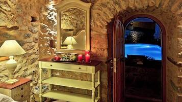 A room with a stone wall, a wooden door leading to a pool, a bedside table with a lamp, and a mirror.