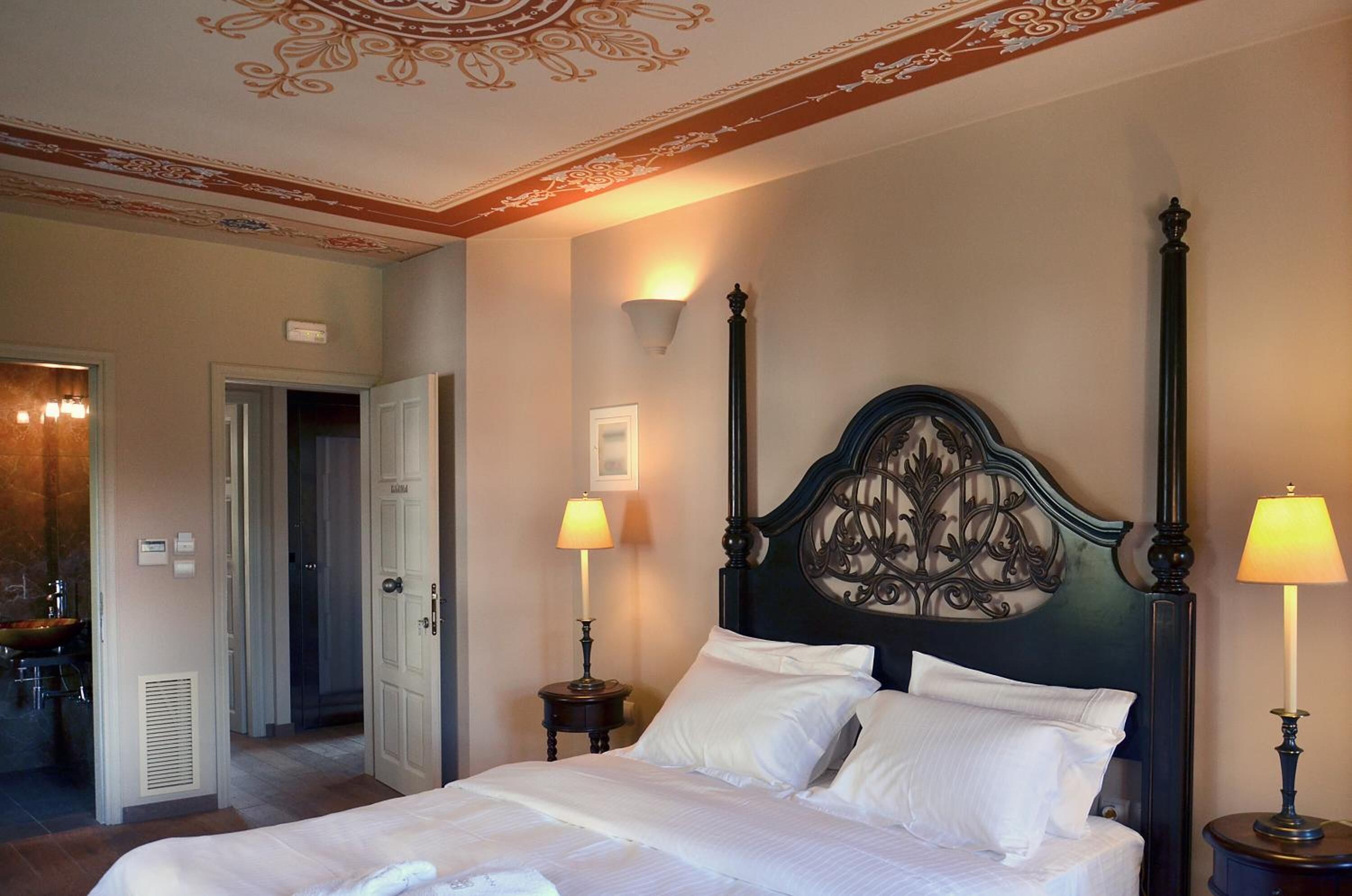 A hotel room with a large bed, two bedside lamps, a decorative headboard, and a bathroom visible through an open door.