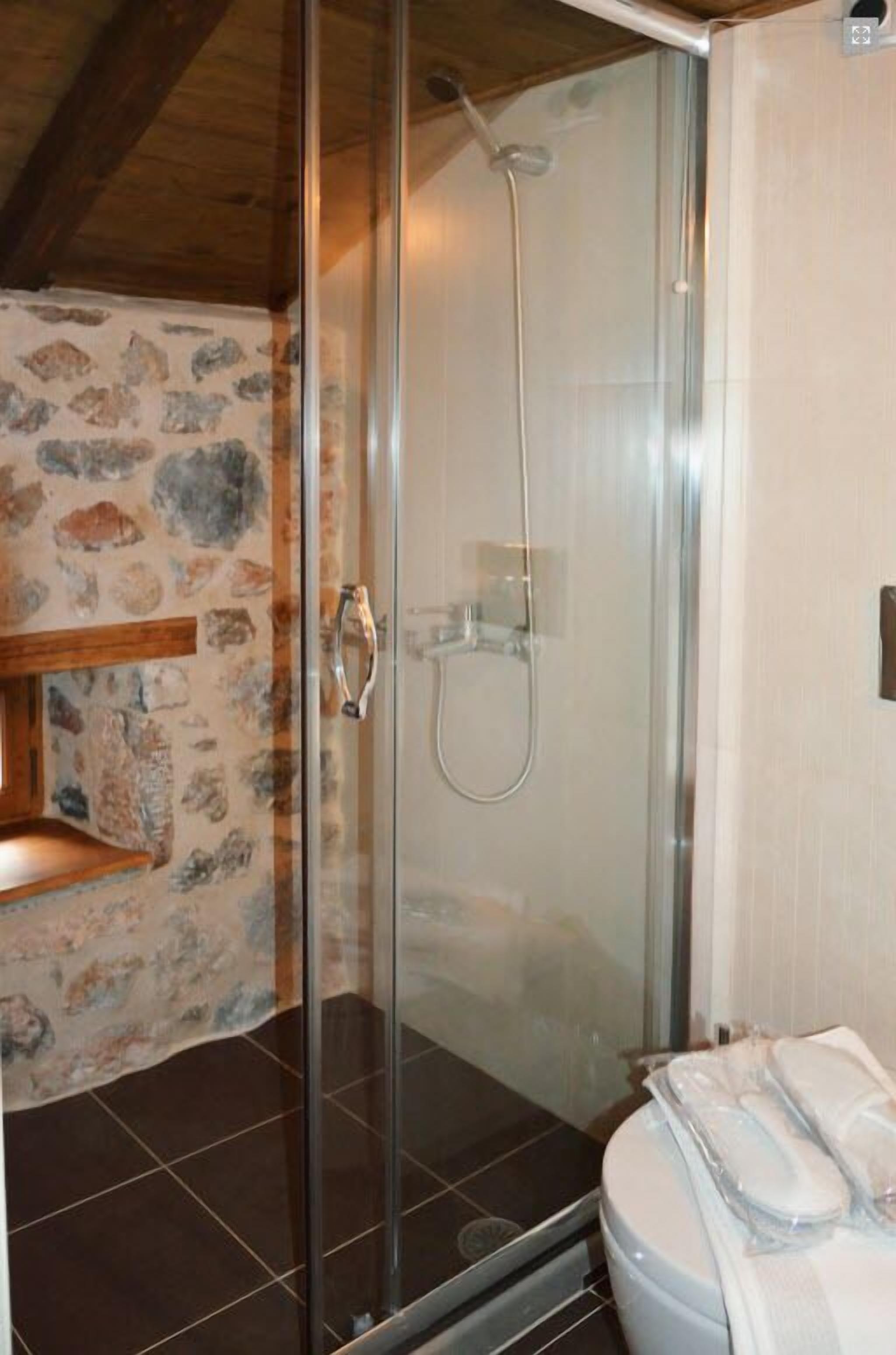 Une salle de bain avec une douche à parois de verre, un mur en pierre, une étagère en bois et des toilettes avec une serviette.