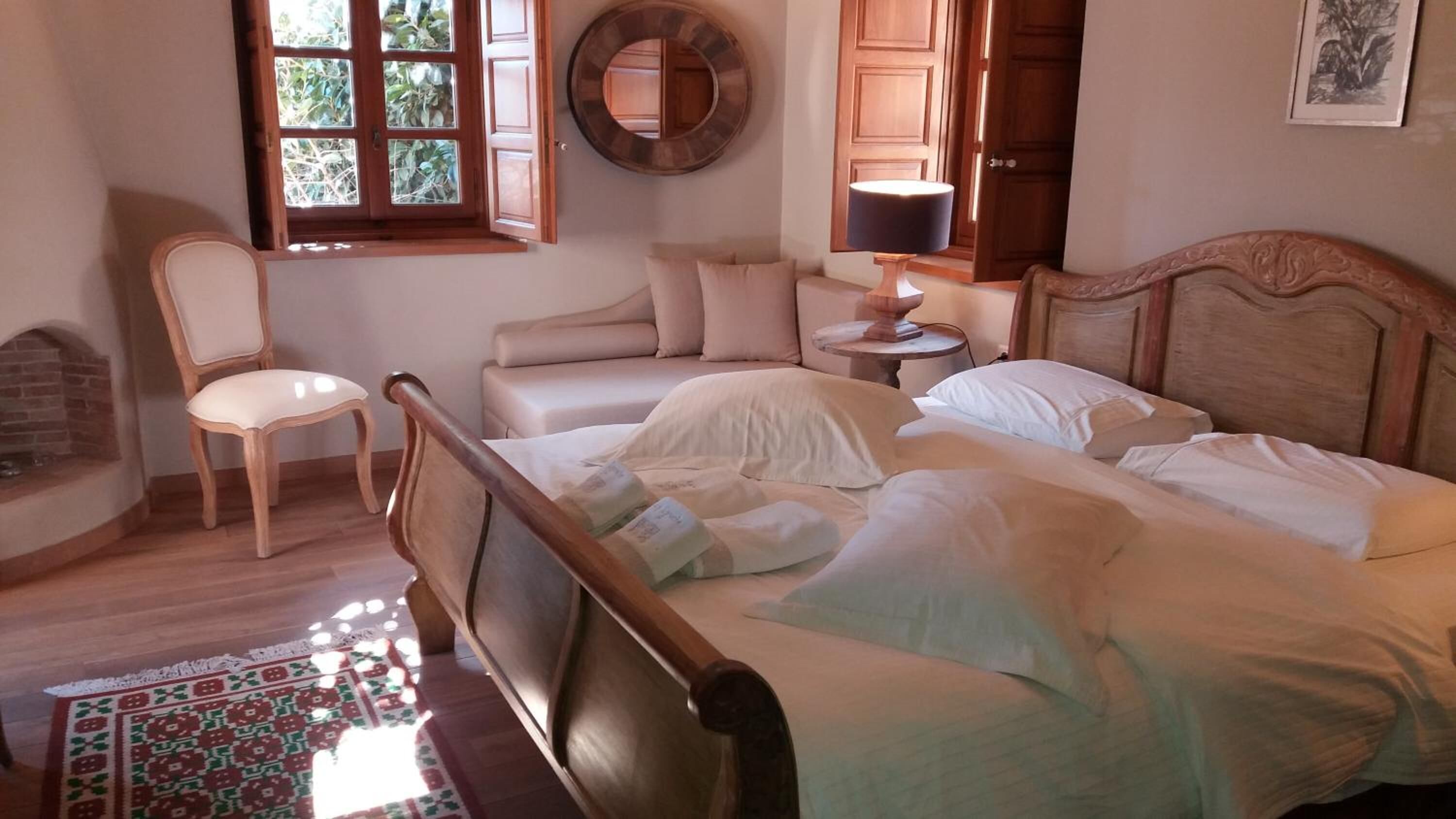 A bedroom with a wooden bed, a chair, a bedside table with a lamp, a fireplace, and a window with a view of greenery.